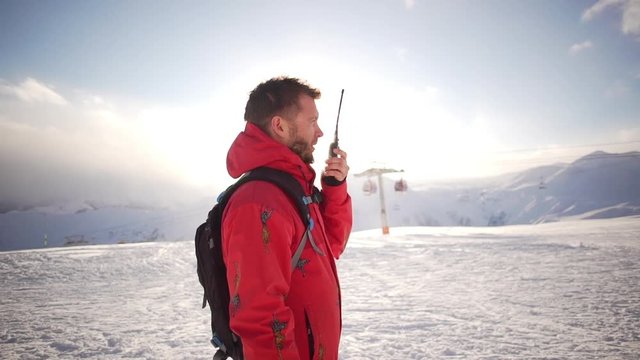 Caucasian male with beard and red parka standing talking on radio on ski mountain resort in slowmotion. Poma lift on background