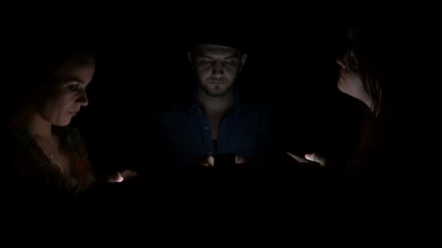Two young women and man in the dark using their smart phones to socialize