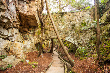 Fototapeta premium A stone bridge over a pedestrian footpath beside a rock cliff dotted with trees and green foliage