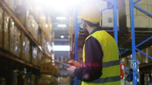 Surveyor Wearing Hard Hat with Clipboard Fills in Forms in a Warehouse. He Walks Through Rows of Storage Racks with Merchandise. Shot on RED EPIC-W 8K Helium Cinema Camera.
