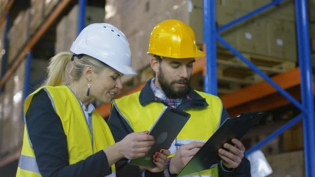 Female Supervisor with Tablet Computer and Male Surveyor with Clipboard Have Discussion in Warehouse with Big Pallet Racks in it. Shot on RED EPIC-W 8K Helium Cinema Camera.