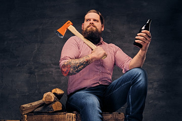 Bearded lumberjack male with tattoos on arms holds the axe and beer bottle