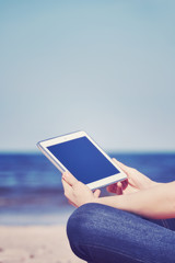 Color toned close up of female hands with tablet computer on a beach, blank screen with copy space, selective focus.