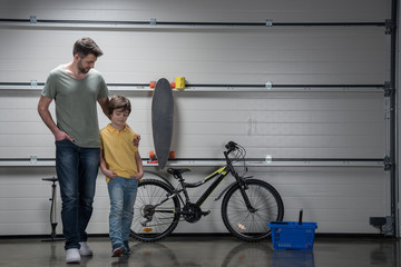 Smiling father and son standing together in workshop with bicycle and skateboard