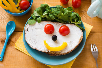 Kids meal, breakfast or lunch for children. Colorful funny food face with healthy vegetables, cheese spread and green salad on a plate. Top view, closeup