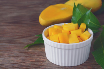 sliced of ripe yellow mangoes with fruit and green mango leaf served with white bowl on wood table in the background.