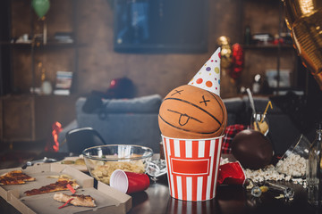 Close-up view of basketball ball with funny face in popcorn box and messy table after party
