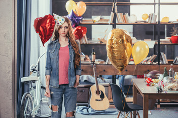 Upset young woman in party hat standing in messy room after party