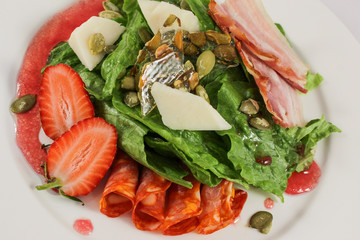 Mix of hot sausages with bacon on a cushion of lettuce leaves, in a duet with parmesan and strawberries, garnished with pumpkin seeds, dressed with raspberry sauce