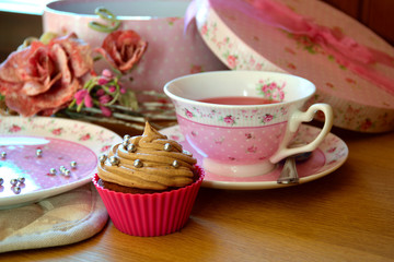 Pink chocolate cupcake frosted with chocolate butter-cream, pink teacup and plate, roses and pink gift box, afternoon tea