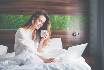 Pretty woman on bed in modern apartment using laptop and drinking coffee after wake up