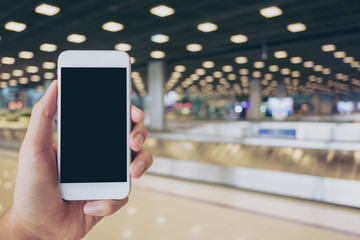 Mockup image of a man holding white mobile phone with blank black screen while standing and waiting for baggage claim in the airport