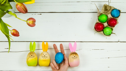 Bouquet of tulips, painted Easter eggs and 3 wooden bunnies with a hand in between holding a painted egg while making a bunny ears gesture on white wooden background