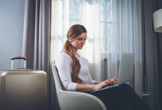 Pretty Woman In Modern Hotel Room Sitting On Armchair And Using Laptop