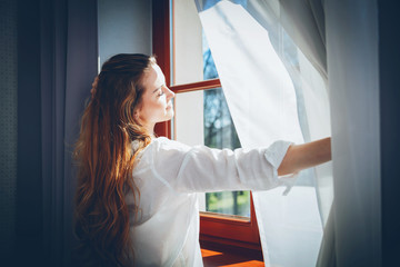 Pretty woman in modern apartment opening window curtains after wake up