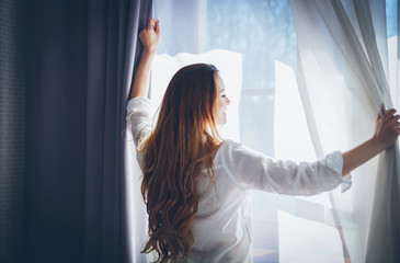 Pretty woman in modern apartment opening window curtains after wake up