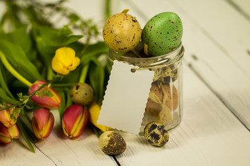 Bouquet of different colored tulips, a jar full of quail eggs and colored Easter eggs with empty label on white wooden background 