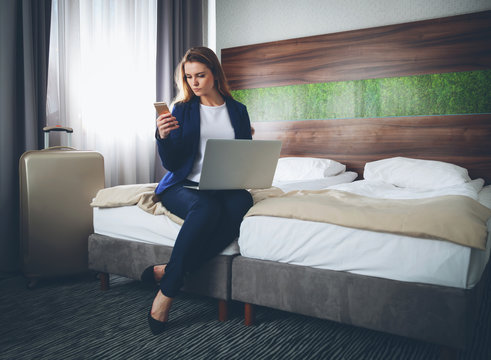 Business woman with suitcase in modern hotel room using laptop and smartphone