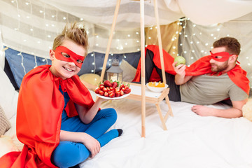 Happy father and son in superhero costumes eating fruits in blanket fort