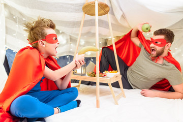 Fototapeta na wymiar Excited father and son in superhero costumes having fun with fruits in blanket fort