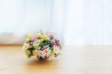 Colorful of artificial flowers in a ceramic pot on wood table near window with curtain.