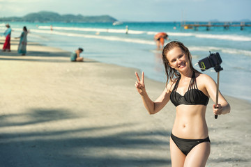 Young woman taking selfie with smartphone. Sexy girl in black bikini on beach taking selfie using monopod