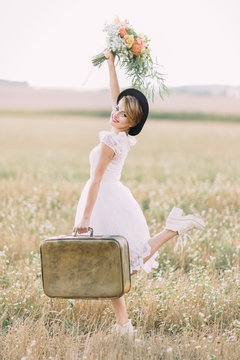 The full-length portrait of the stylish bride carrying the vintage suitcaise and keeping up the colourful bouquet in the field. The side view.