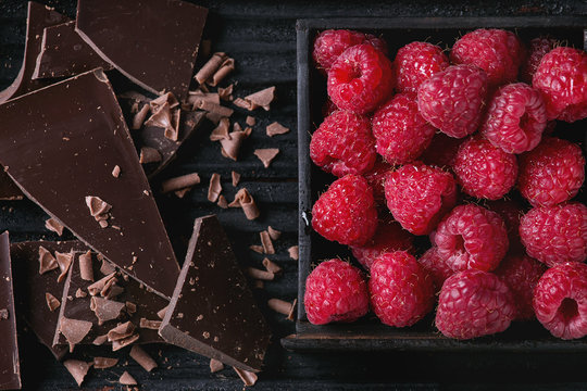 Dark and milk chopping chocolate and chips with fresh raspberries in black wood box over black burnt wooden background. Top view, close up. Chocolate dessert concept