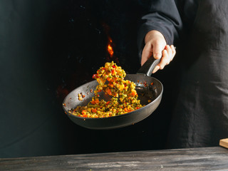 Cooking of vegetable mix stew with tossing on a hot frying pan in the tongues of flame. Colorful grains of corn and peas, asparagus and sweet peppers.