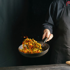 Cooking of vegetable mix stew with tossing on a hot frying pan in the tongues of flame. Colorful grains of corn and peas, asparagus and sweet peppers.