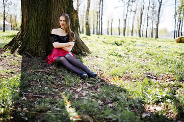 Portrait of girl with bright make up with red lips, black choker necklace on her neck and red leather skirt sitting near tree at park.