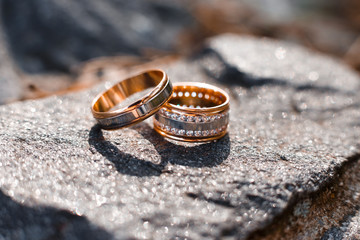 Gold wedding rings with a lot of gemstones lying on the grey stone and shining in the sun rays. Jewelry made of white and yellow gold