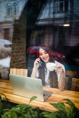 Woman at the cafeteria using phone and laptop and drink cup of coffee