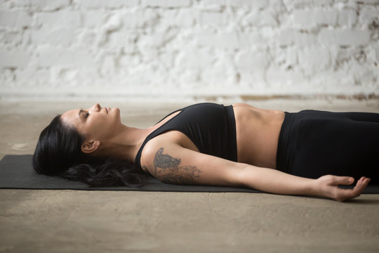 Middle aged yogi woman practicing yoga concept, lying on floor in Savasana exercise, Corpse, Dead Body pose, working out, wearing sportswear, black top and pants, close up, white loft background