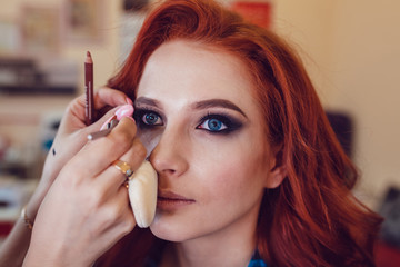 Make-up artist does makeup to the red-haired girl in studio. Smoky eyes.