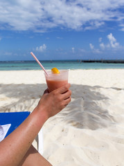 Drink on the beach. A woman is holding a colorful drink. A slice of lemon and a straw is on the drink. White sand beach. Turquoise water in the background.