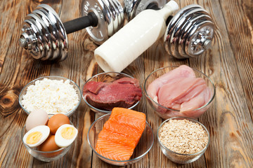 Protein, fish, cheese, eggs, meat, chicken and dumbbells on a wooden background