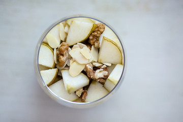 Oatmeal porridge flavored with mashed banana, topped with sliced pear, walnuts, almonds and honey, view from above, served in a clear round glass, placed on a wooden light colored background