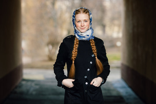 Young beautiful fashionable redhead woman with braids hairdo in blue white headcraft stylish denim black trench jacket posing outdoors in street arc spring time
