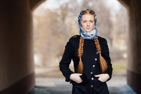 Young beautiful fashionable redhead woman with braids hairdo in blue white headcraft stylish denim black trench jacket posing outdoors in street arc spring time