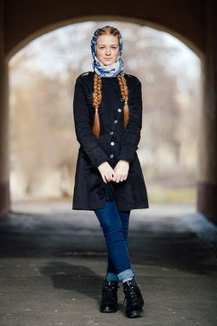 Young beautiful fashionable redhead woman with braids hairdo in blue white headcraft stylish denim black trench jacket posing outdoors in street arc spring time