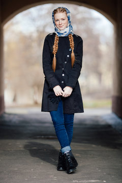 Young beautiful fashionable redhead woman with braids hairdo in blue white headcraft stylish denim black trench jacket posing outdoors in street arc spring time
