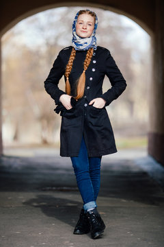 Young beautiful fashionable redhead woman with braids hairdo in blue white headcraft stylish denim black trench jacket posing outdoors in street arc spring time