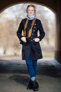 Young beautiful fashionable redhead woman with braids hairdo in blue white headcraft stylish denim black trench jacket posing outdoors in street arc spring time