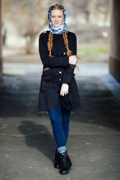 Young beautiful fashionable redhead woman with braids hairdo in blue white headcraft stylish denim black trench jacket posing outdoors in street arc spring time