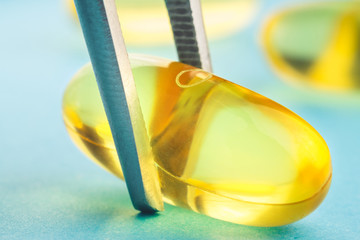 Tweezers keeps one yellow capsule of omega 3, fatty acid, fish oil on blue background, macro image.