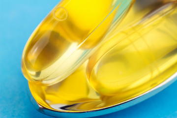 Two yellow capsules of omega 3, fatty acid, fish oil in the spoon, on blue background, macro image.