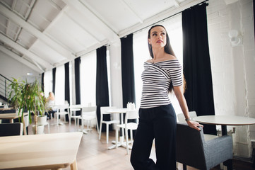 Obraz premium Beautiful brunette girl standing in cafe near wooden table. Well-groomed woman sitting in modern interior with lights background looking away.