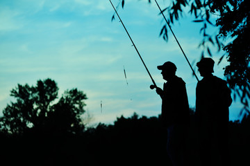 Silhouettes of two fishermen with fishing rods on nature background and blue sky. Fishing