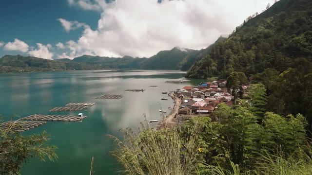 View of traditional Trunyan village on Lake Batur bordered by mountain ridge on sunny summer day with blue sky and clouds. Bamboo fish farms in water. Shot with Sony a7s on tripod in Bali, Indonesia